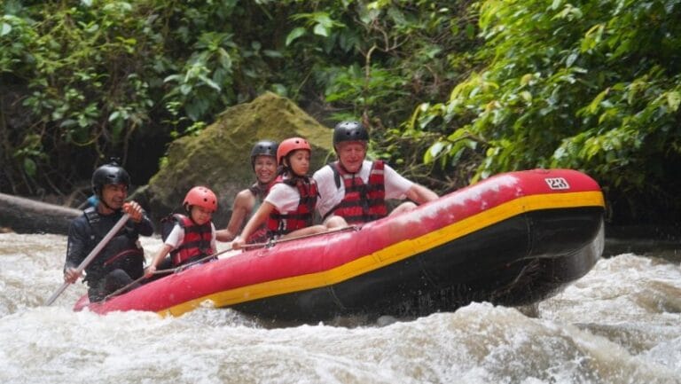 Wisatawan memakai helm dan pelampung sambil mendayung perahu karet di sungai berbatu.